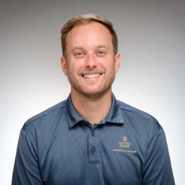 Man with short brown hair and beard smiling in a blue Notre Dame Undergraduate Education polo shirt.