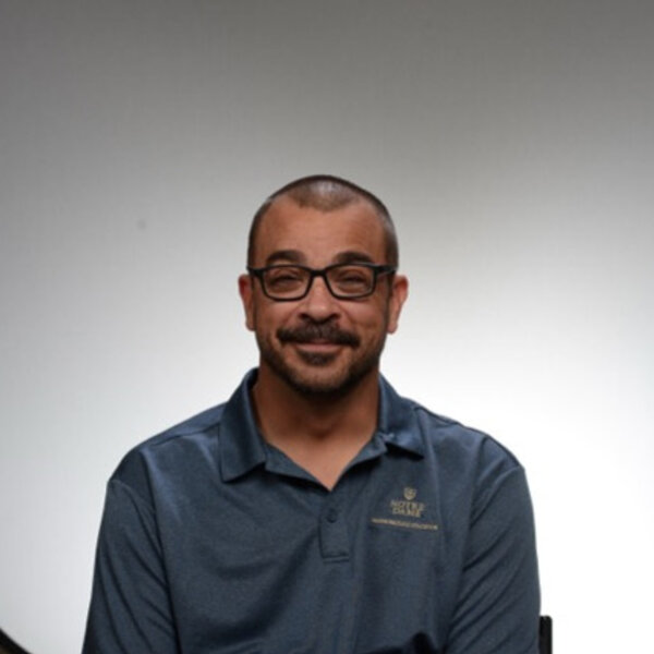 Man with glasses smiles, wearing a blue Notre Dame polo shirt.